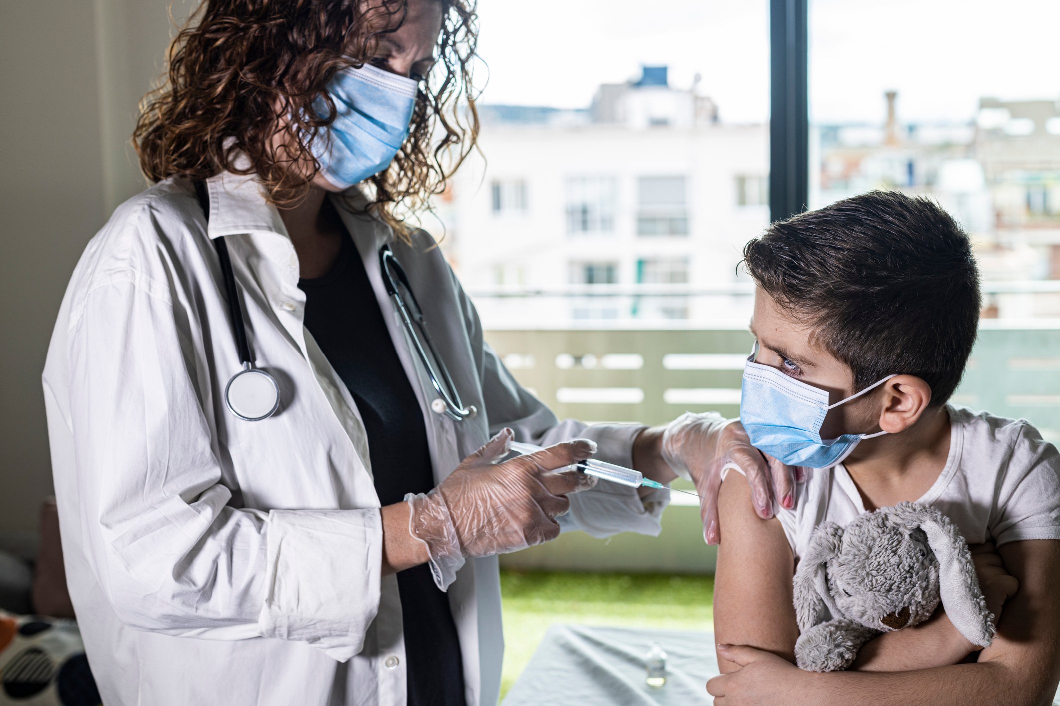 Child holding a stuffed animal getting a shot in the arm from a doctor. 