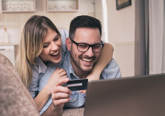 A smiling couple leans over a computer with a credit card while shopping online.