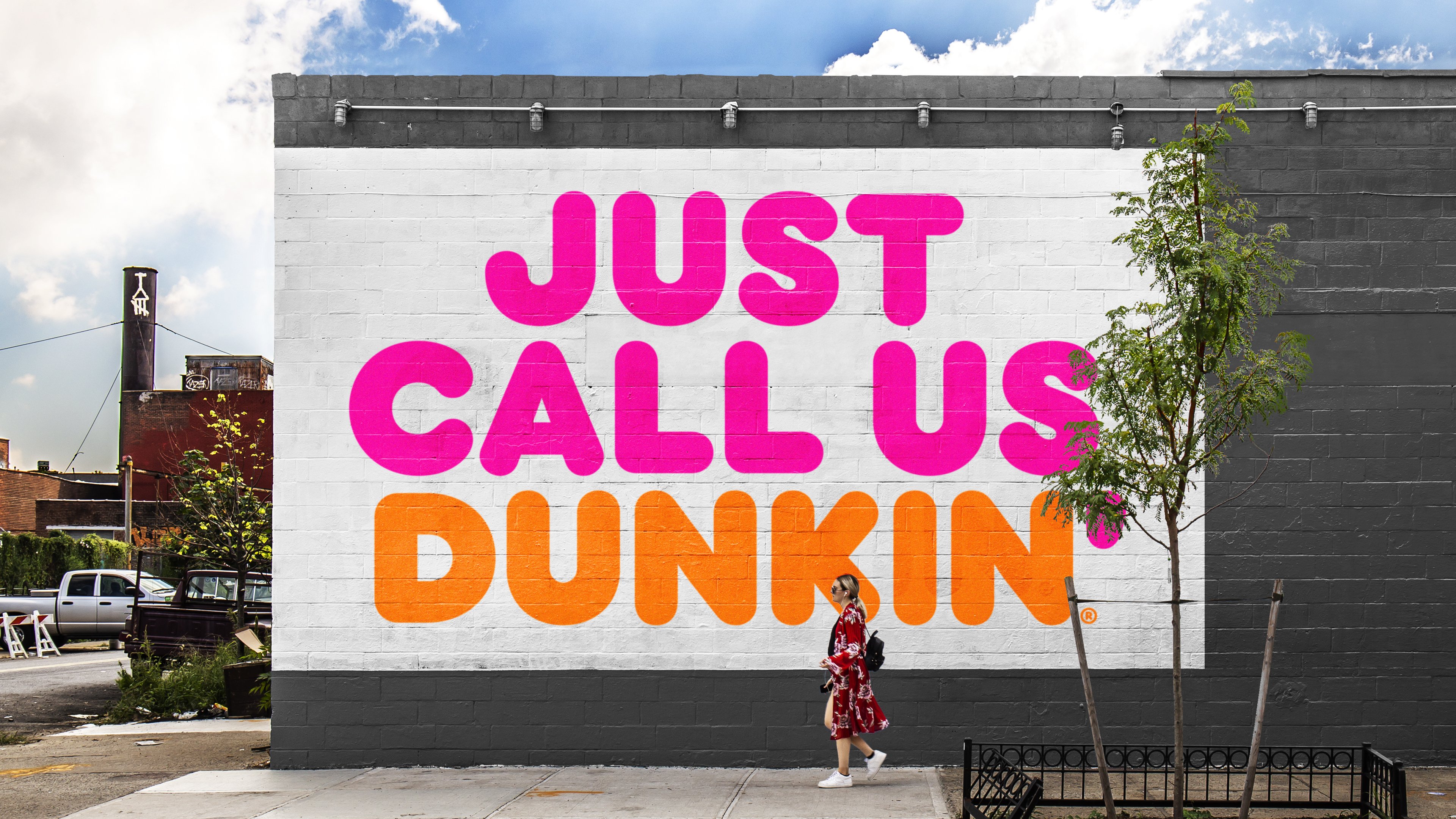 Large sign reading Just Call Us Dunkin painted on a wall, with woman walking past