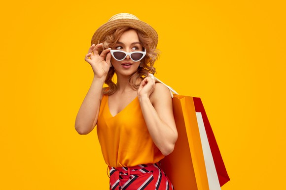 A fashionable woman in a hat and sunglasses with shopping bags slung over  her shoulder. 