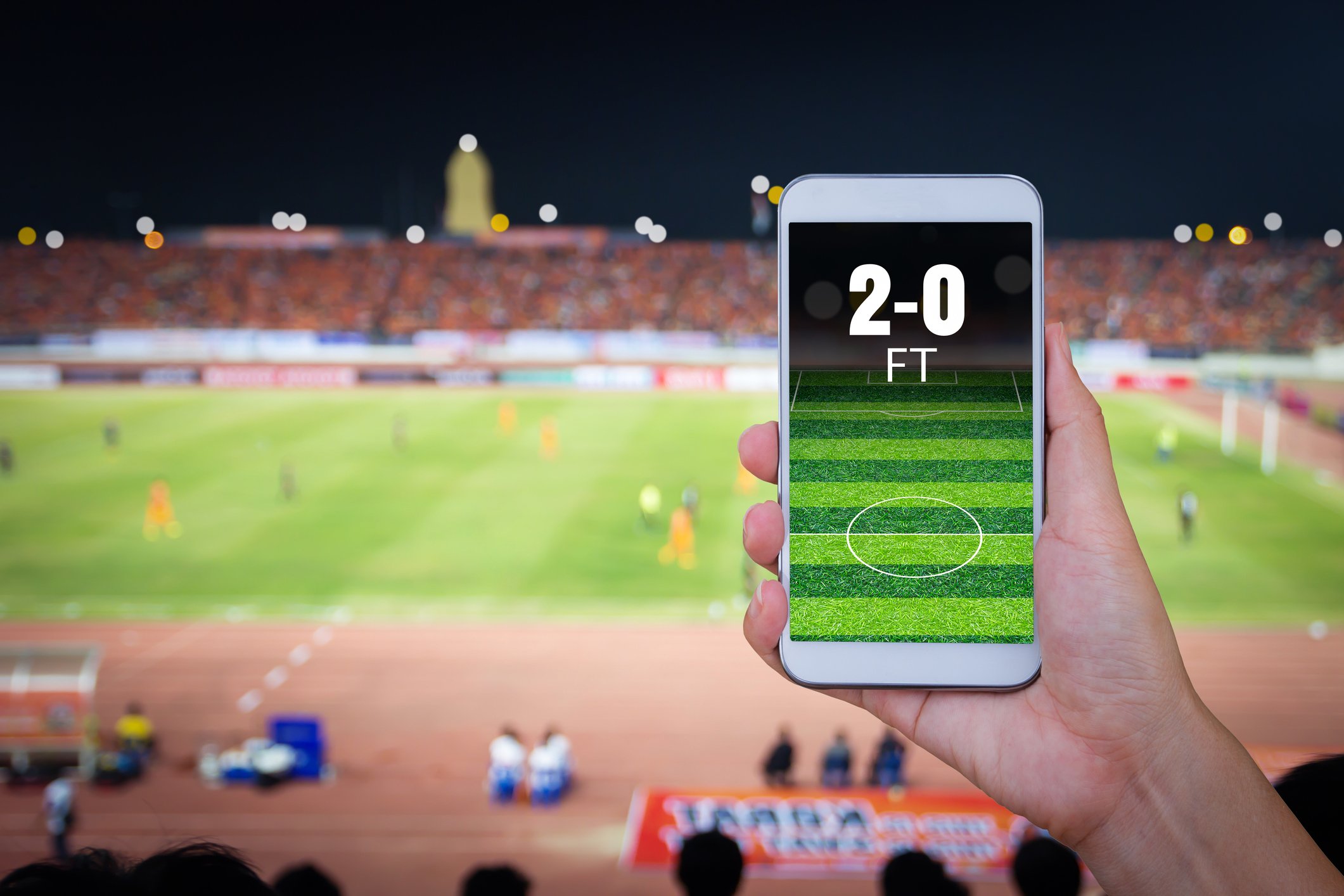 A hand holding up a smartphone with a sports betting app, with a football field in the background.