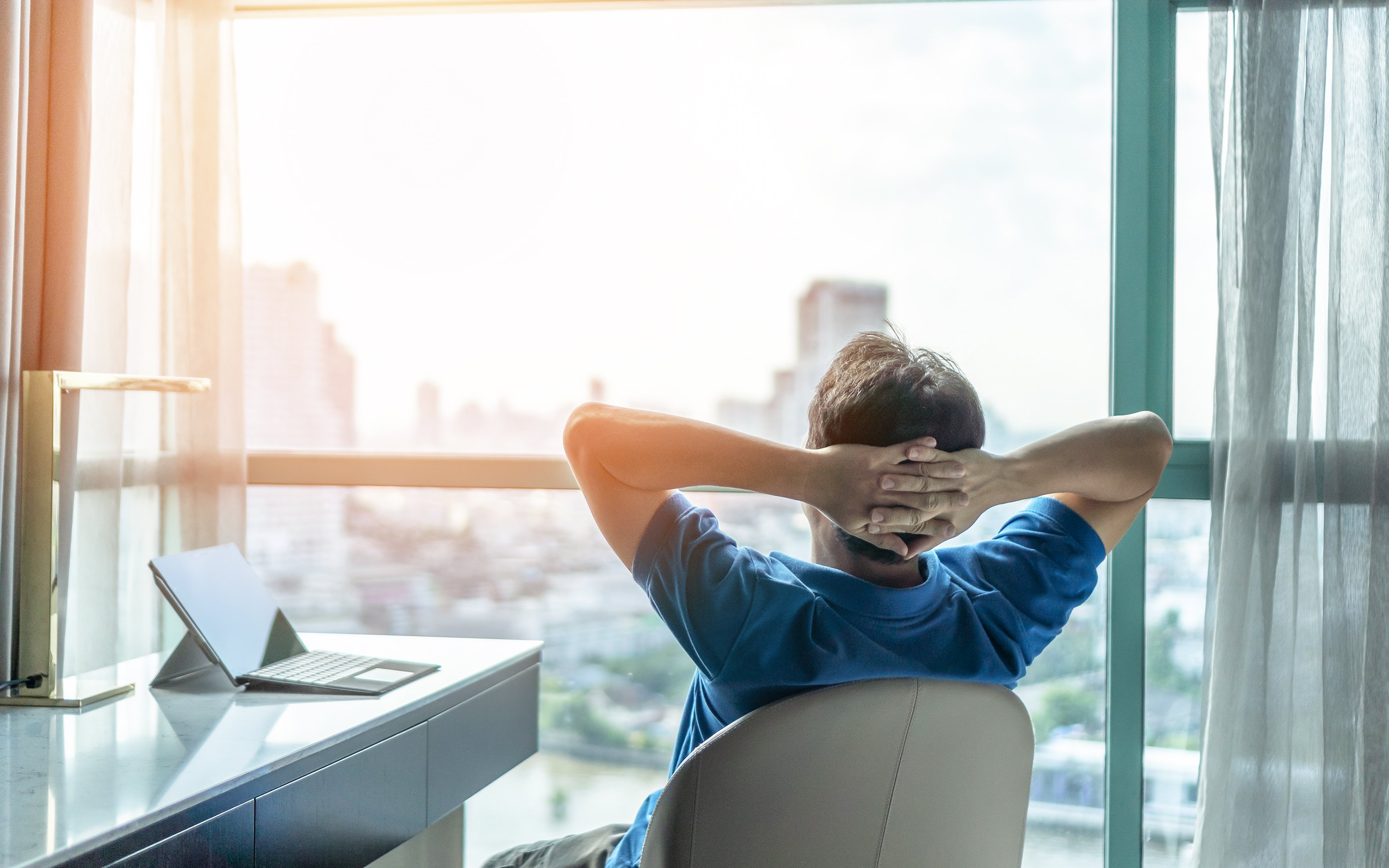 A person leans back in a desk chair with their hands folded behind their head, staring out the window at a city skyline. 