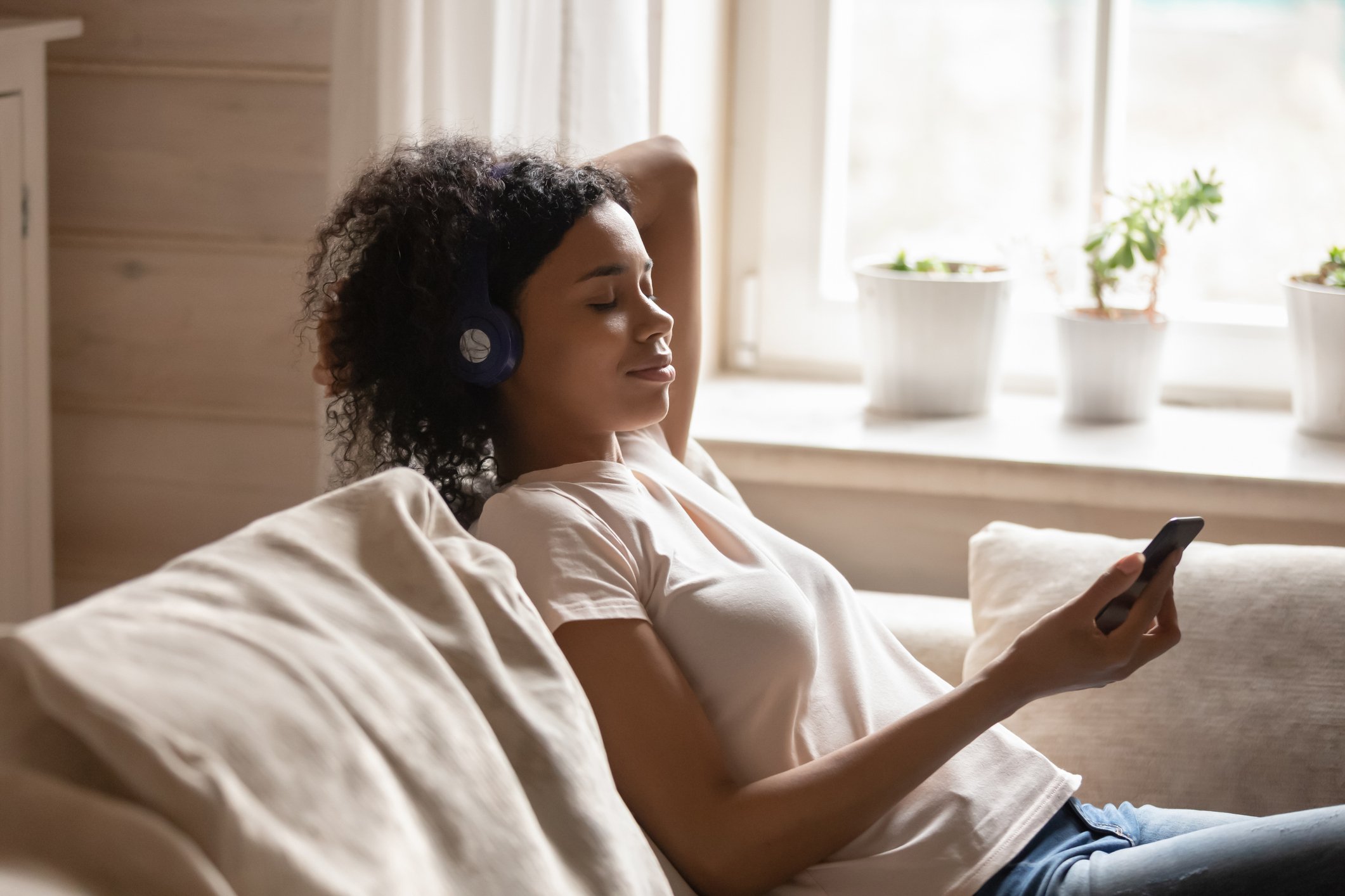 Young woman sitting on couch wearing wireless headphones and looking at her smartphone