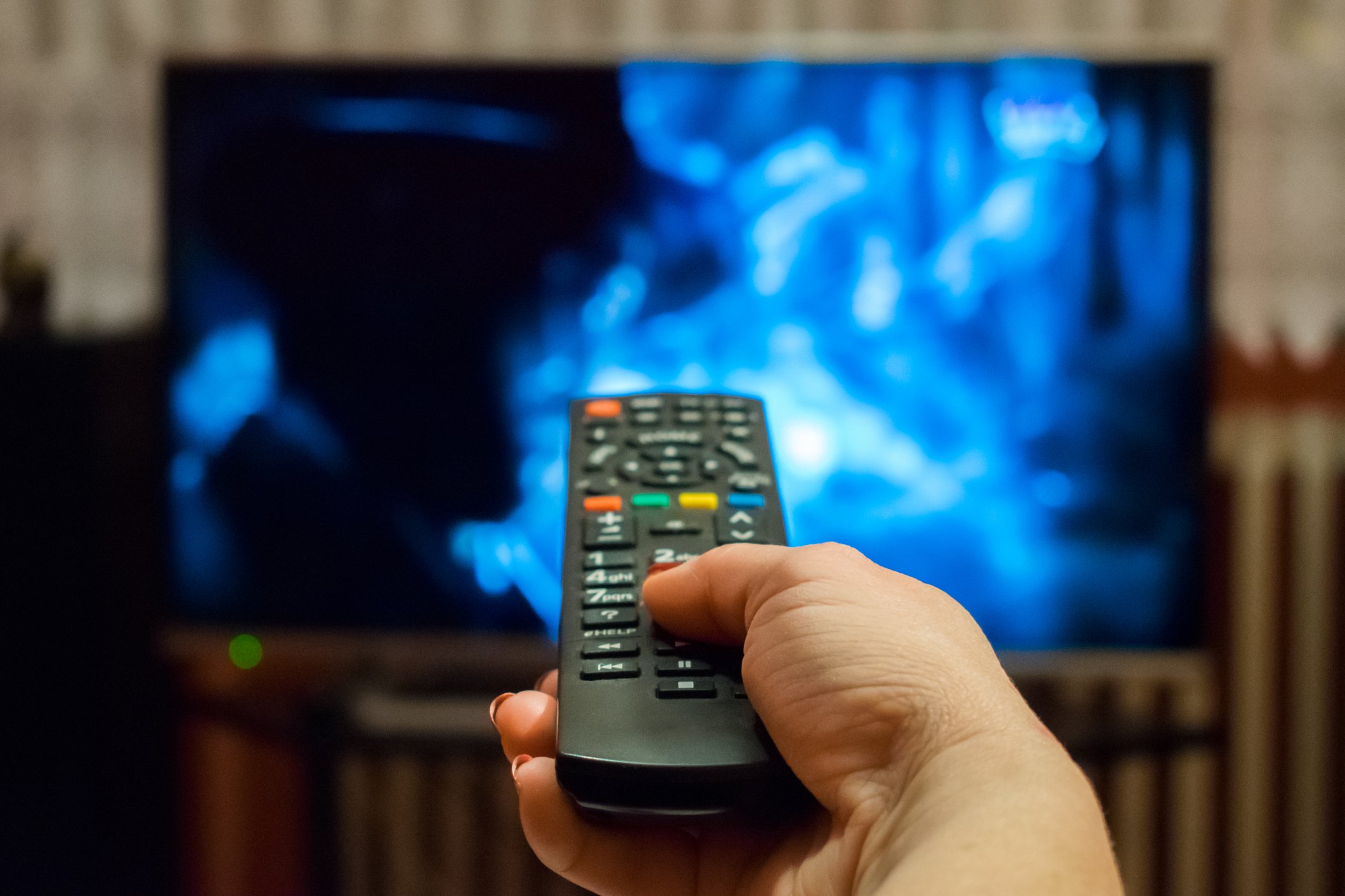 A hand holds a TV remote pointed at a big-screen television set in the background.