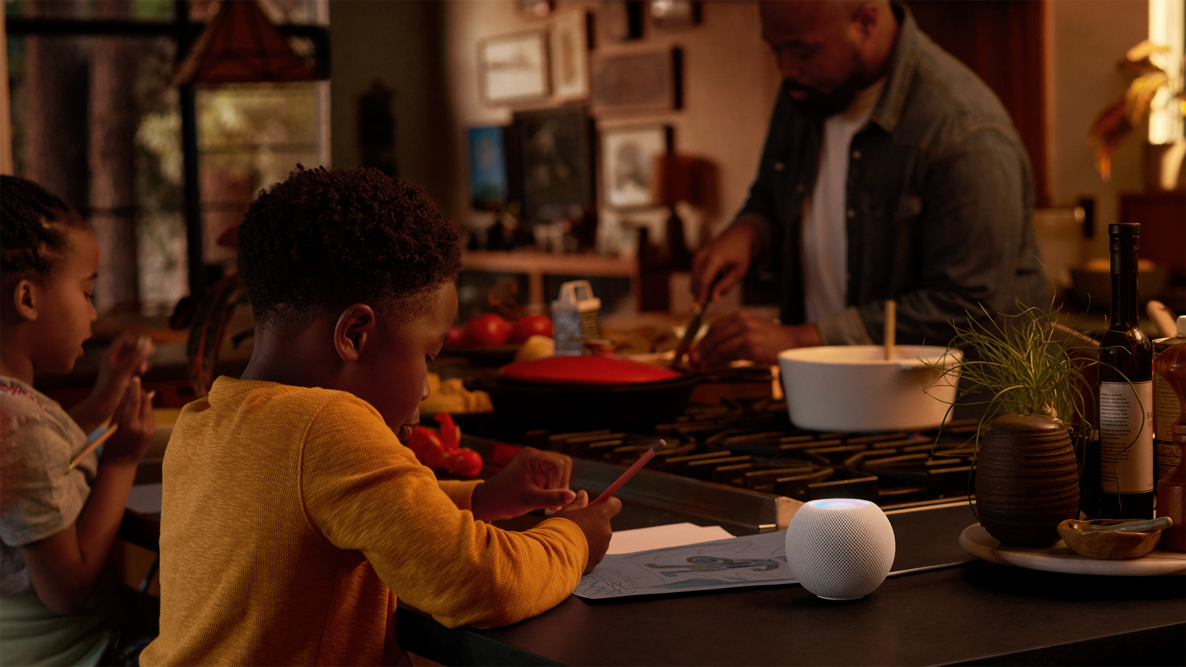 Two children sitting at a kitchen countertop with a man cooking on a stove across from them, with a HomePod Mini foregrounded on the counter