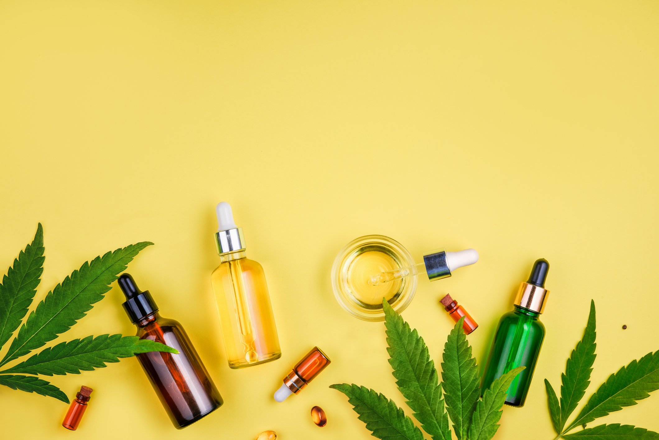 Several glass bottles with cannabis tinctures are scattered on top of a few cannabis leaves.