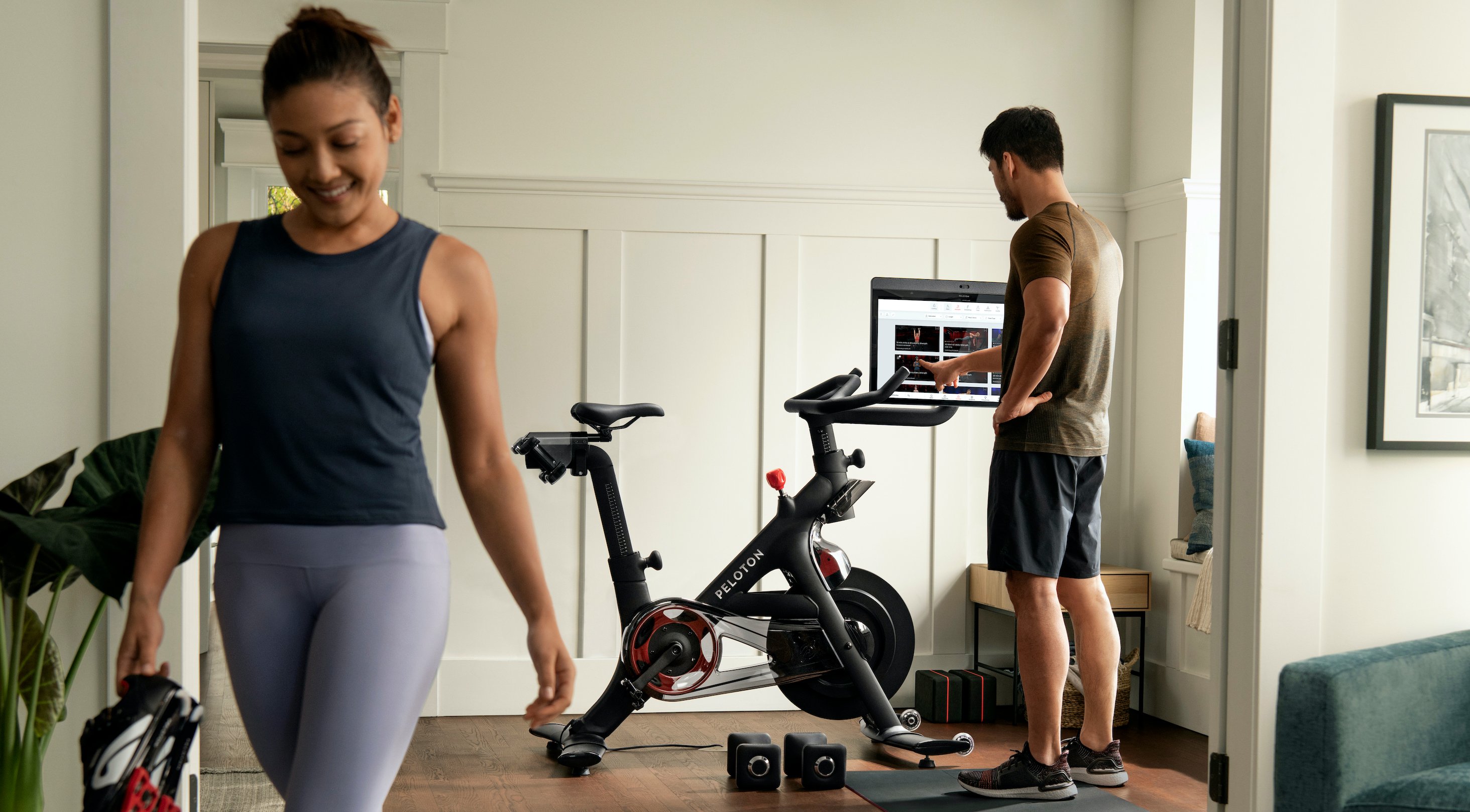 Woman wearing workout clothes walking away from Peloton Bike while man standing next to it reaches towards the screen