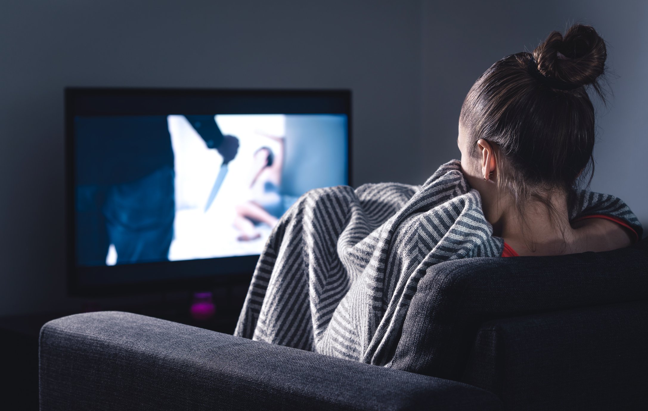 A woman hides beneath a blanket watching a slasher move on TV.