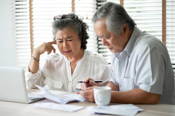 Retirement age man and woman with gray hair looking at financial statements and laptop with concerned expressions.