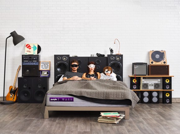 Three people wearing masks lay on a mattress from Purple Innovation in a bedroom setting.