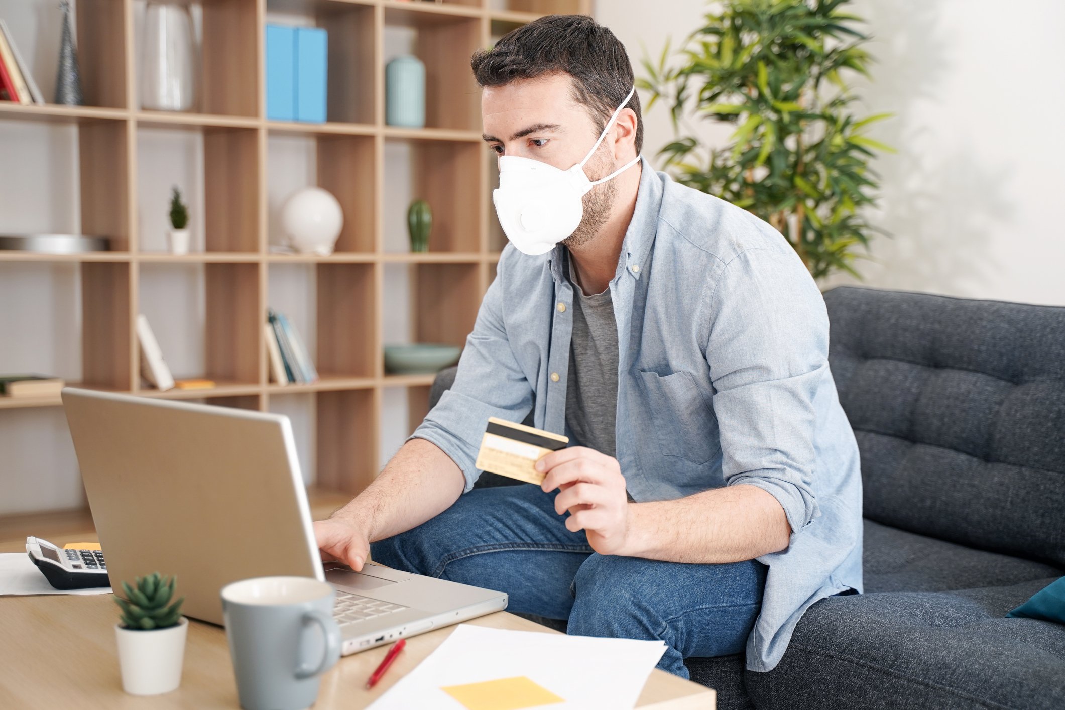 Young man shopping on laptop while wearing a mask and holding a credit card.
