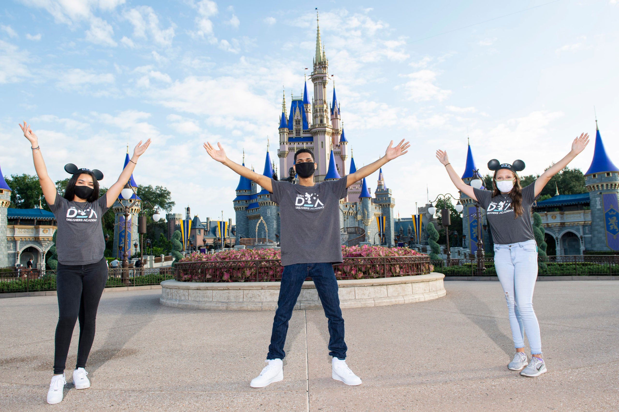 Three young employees wearing masks at Disney World, with the famous castle in the background.