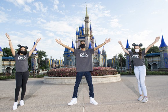 Three young employees wearing masks at Disney World, with the famous castle in the background.