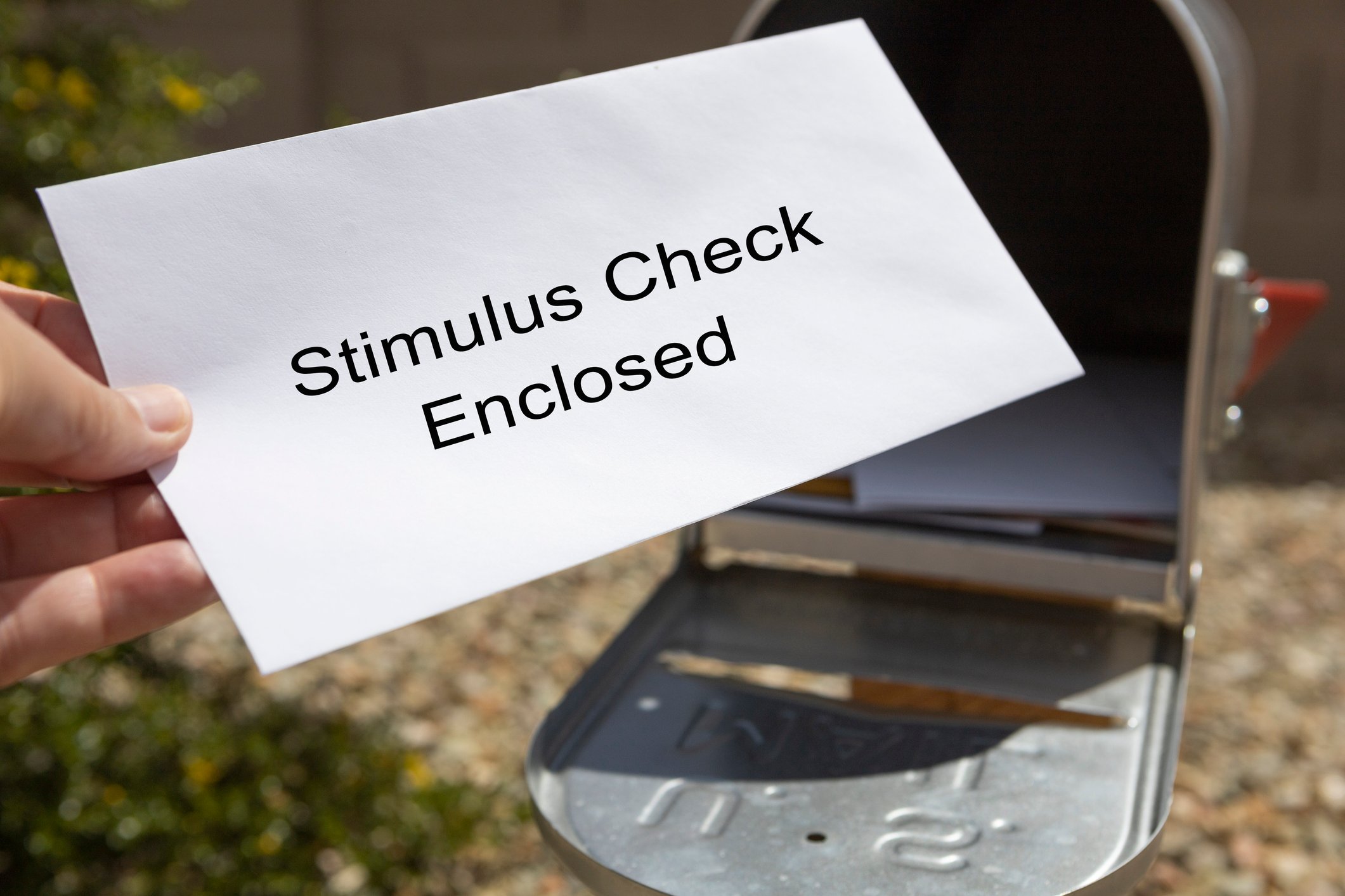 Hand pulling a white envelope out of a mailbox, with the words "stimulus check enclosed" written on the envelope.