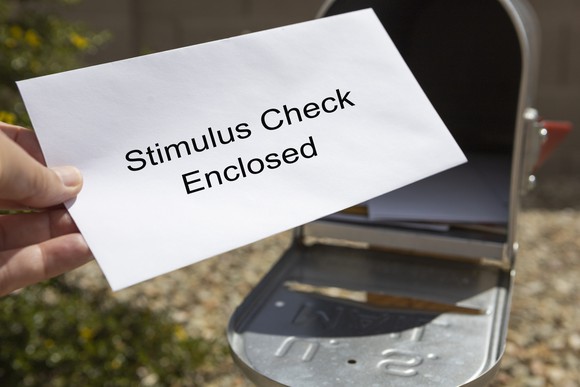 Hand pulling a white envelope out of a mailbox, with the words "stimulus check enclosed" written on the envelope.