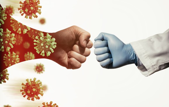 A fist surrounded by images of coronaviruses pushes against a researcher's gloved fist.