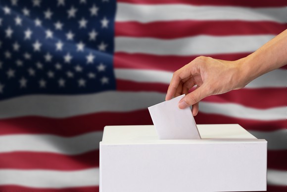 The hand of someone submitting a vote to a ballot box with an American flag in the background.