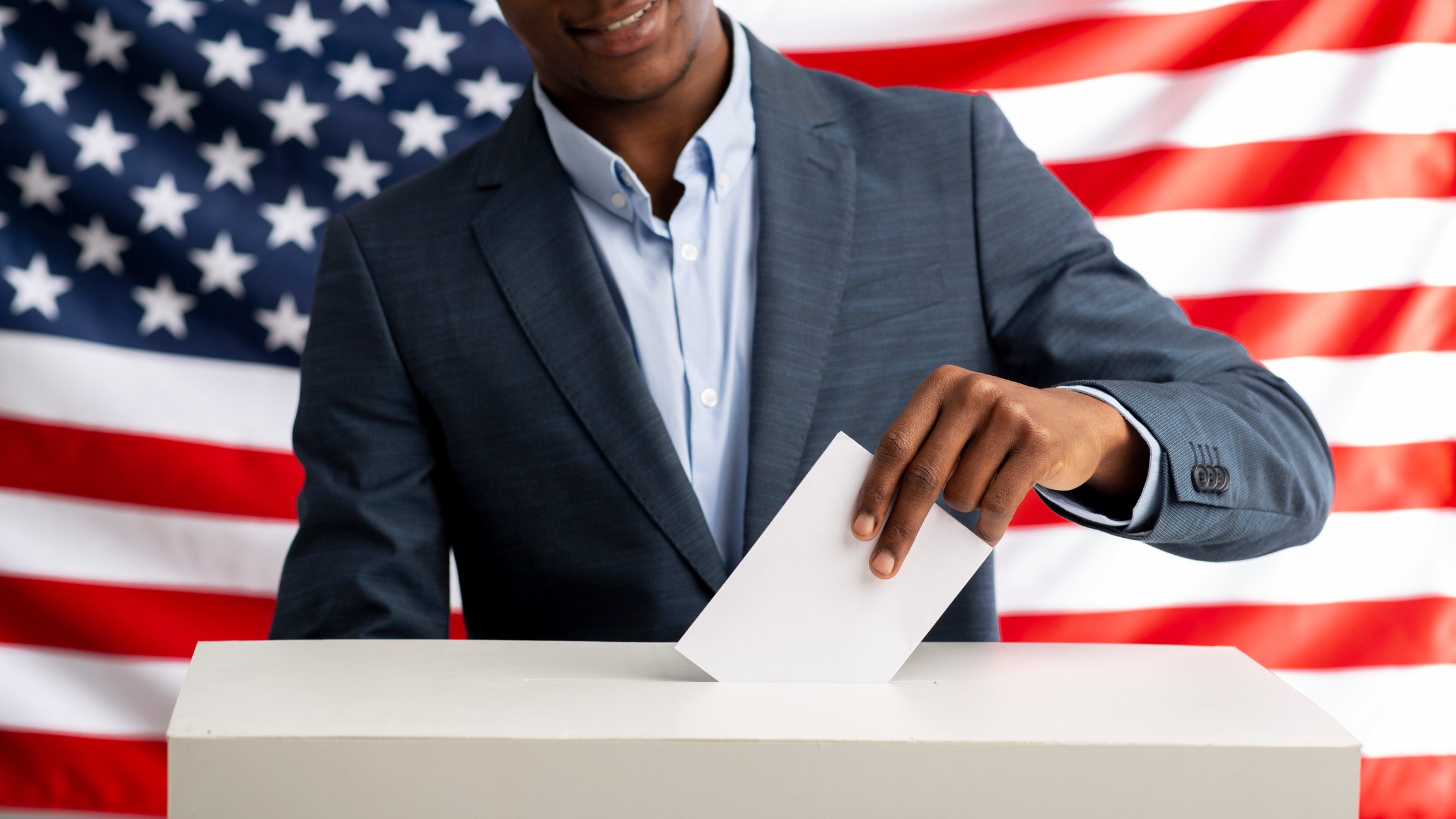 A voter inserts a ballot in the voting box in front of an American flag.