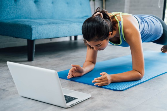 Woman doing push-ups on elbows on yoga mat while looking at a laptop screen.