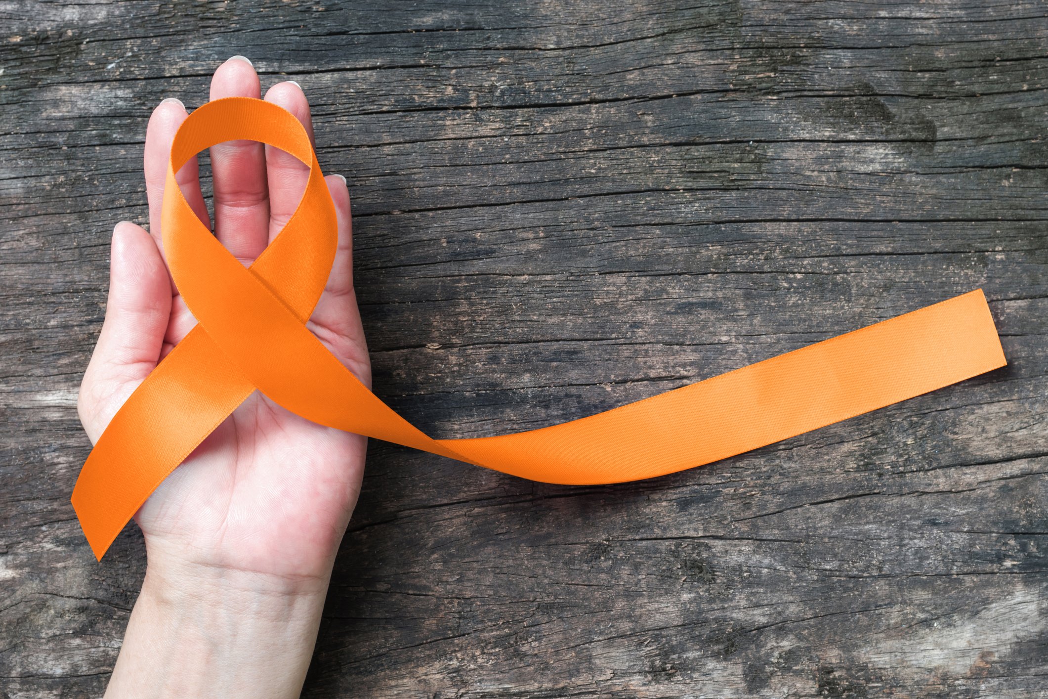 A wood plank background with a hand holding an orange ribbon signifying kidney cancer awareness.