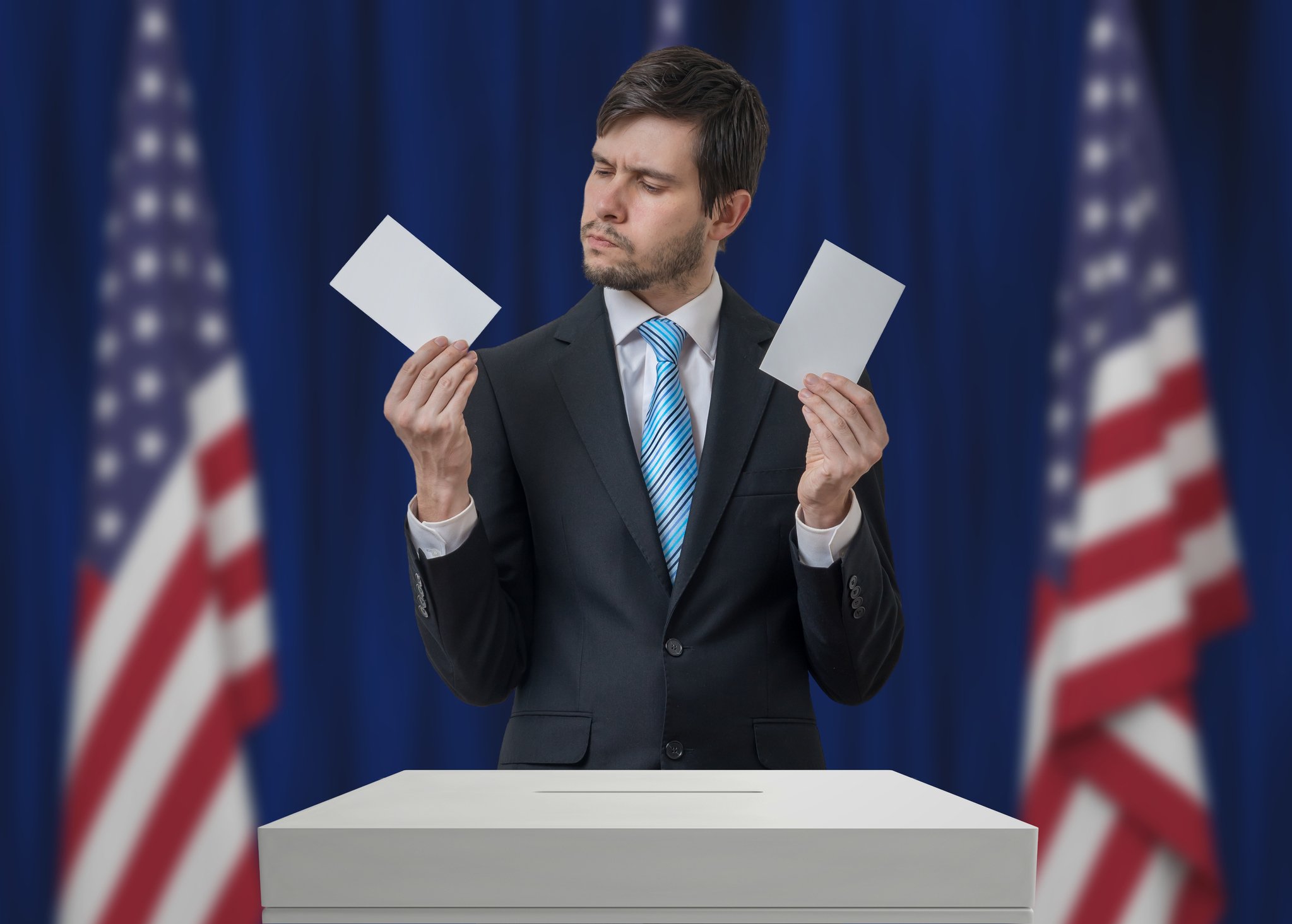A man in a suit at an election box holding two ballots, trying to decide which one to cast.