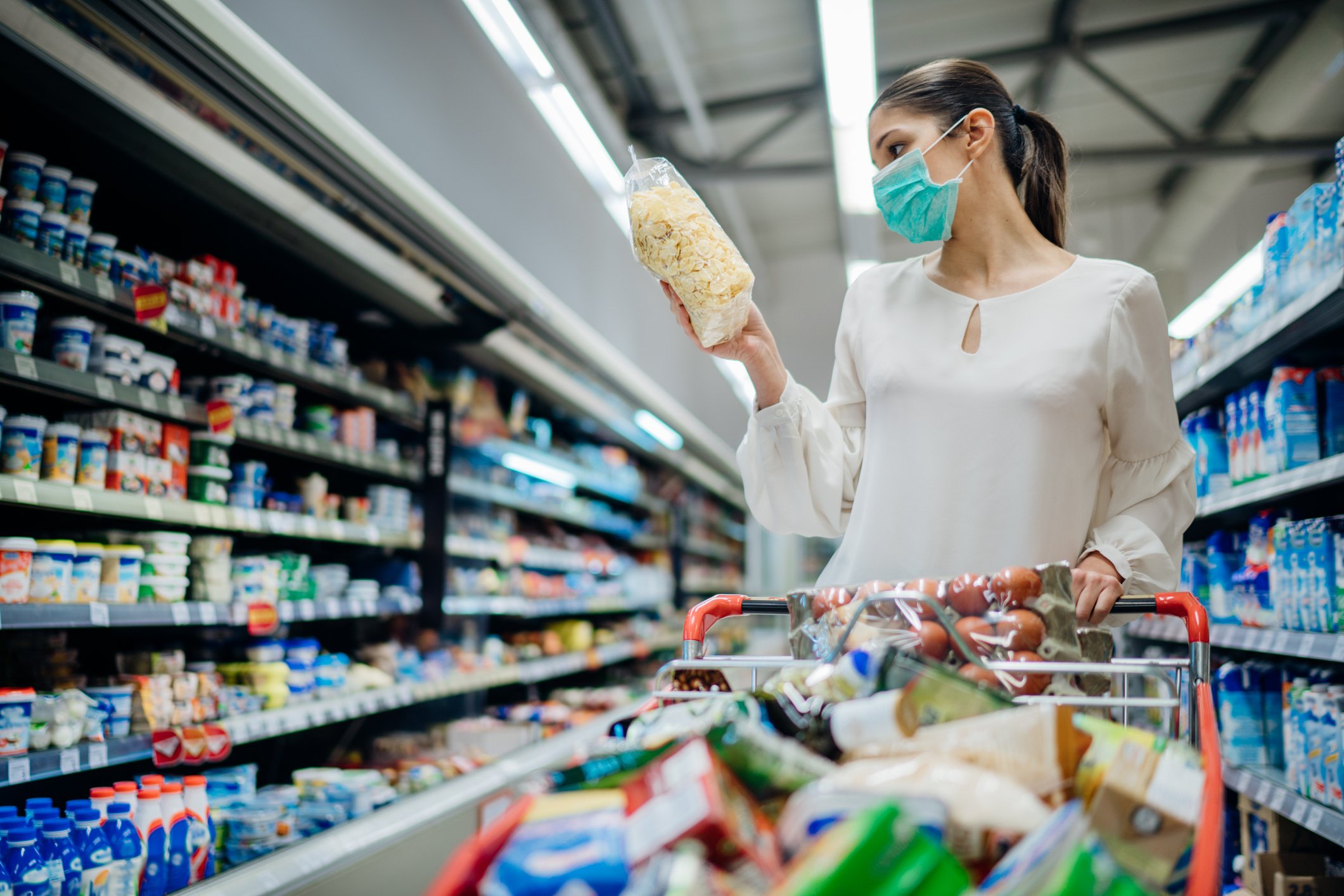 A mask-wearing woman in the aisle of a store. She is holding an item and her cart is full.