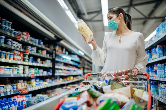 A mask-wearing woman in the aisle of a store. She is holding an item and her cart is full.