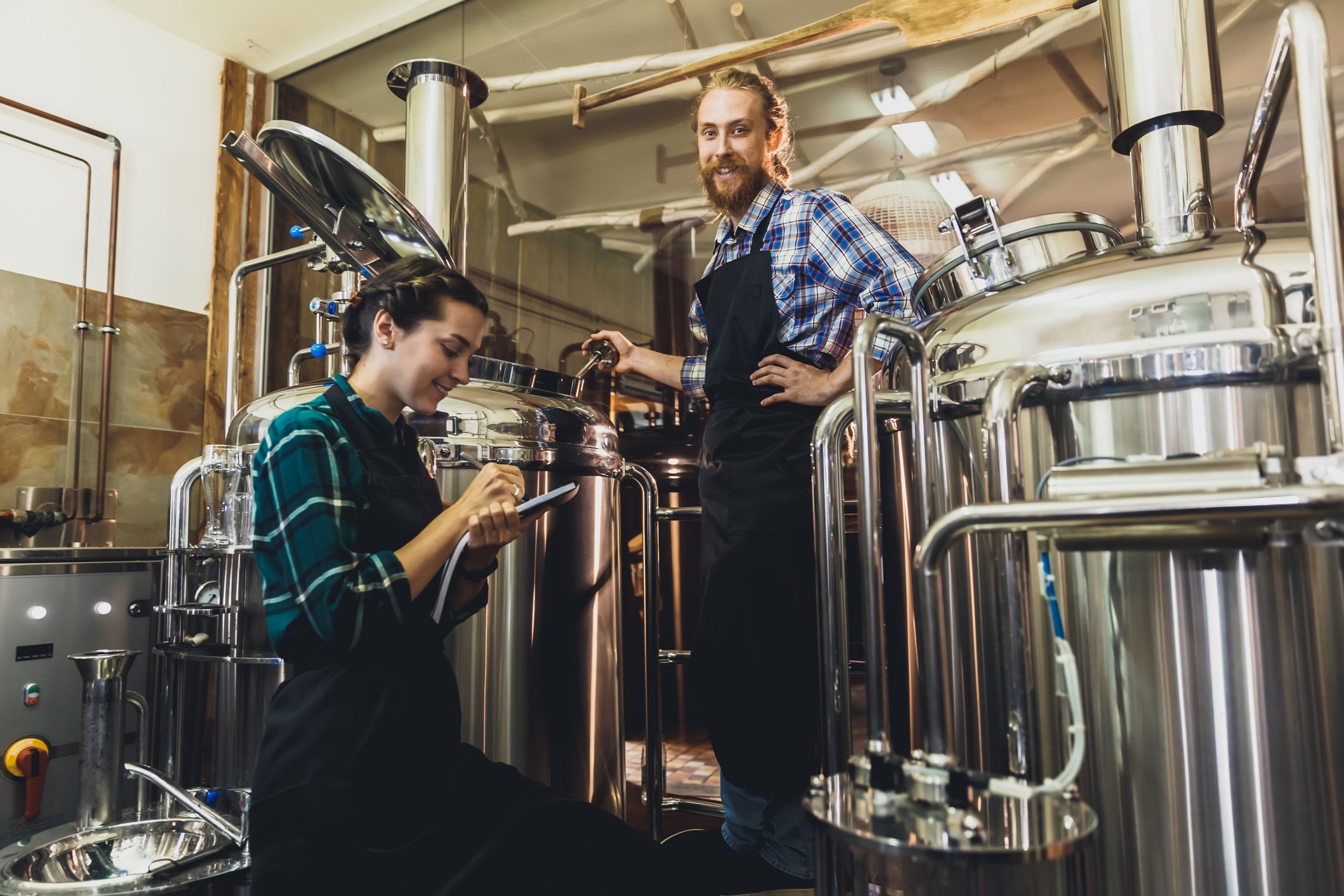 Two workers and stainless steel beer vats and piping at a commercial brewing operation.
