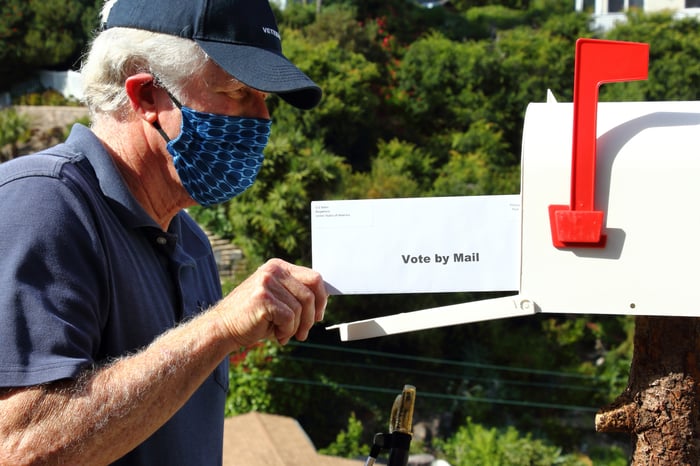 An elderly man with white hair putting an envelope in the mailbox that reads, "vote by mail".