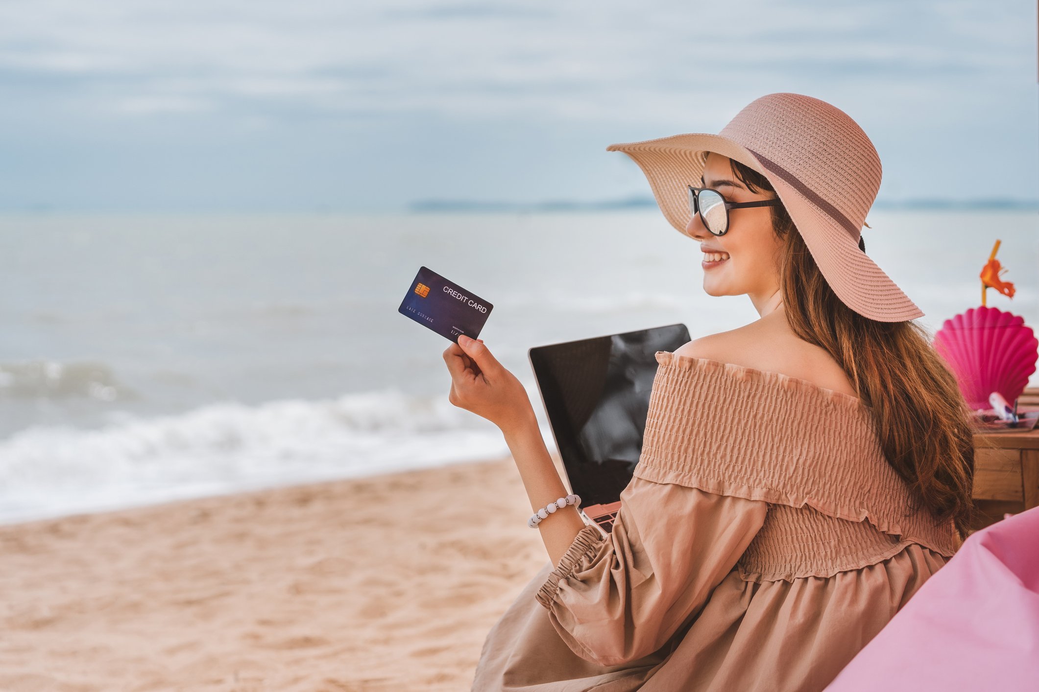 A young woman holds up a card while sitting on a beach with her laptop. 