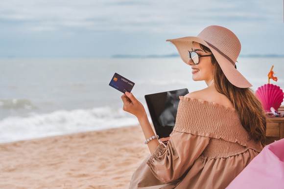 A young woman holds up a card while sitting on a beach with her laptop. 