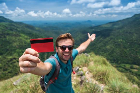 A young man on top of a mountain holds up a credit card. 
