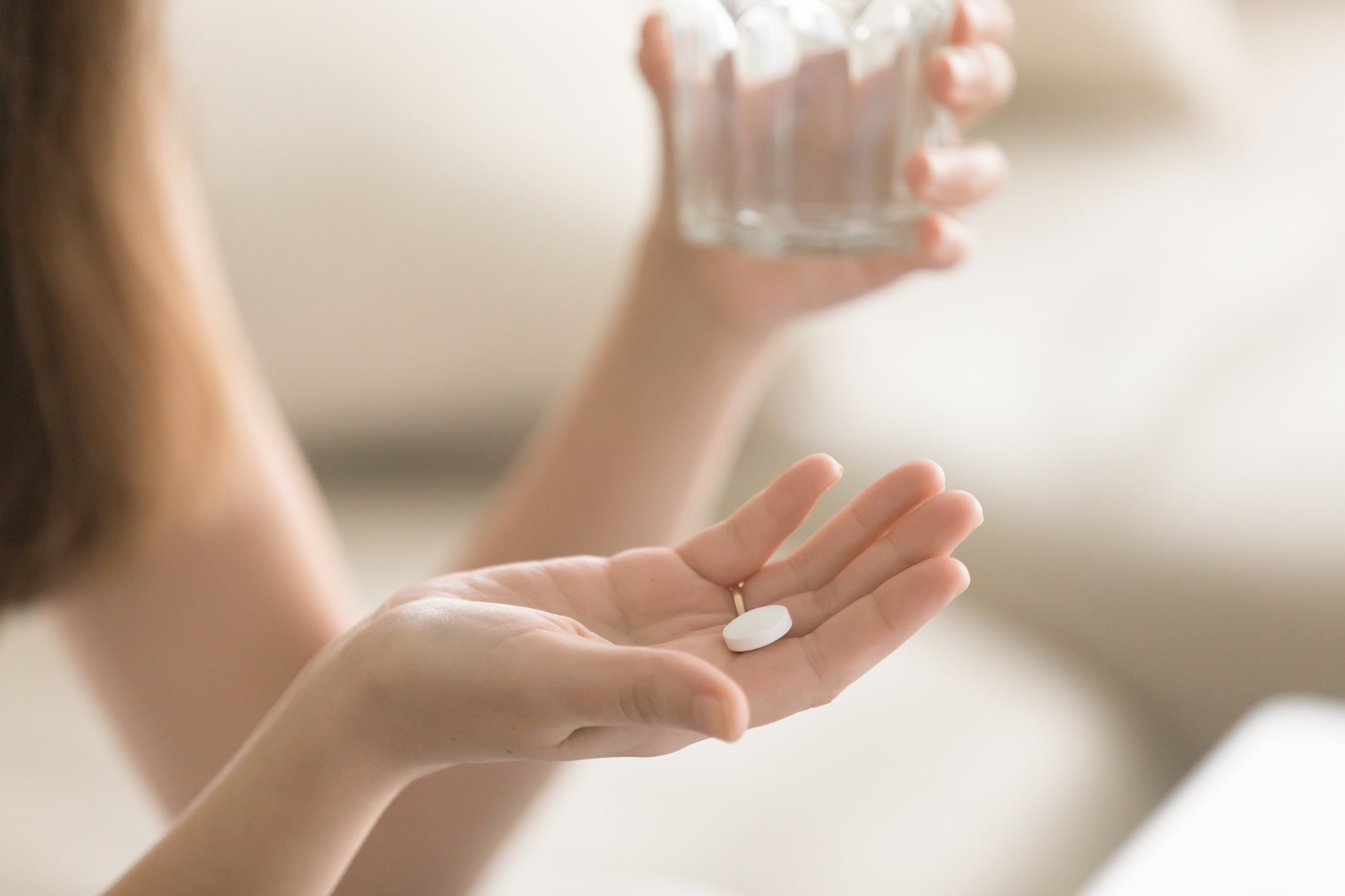 A woman holds a glass in one hand and pill in the other.