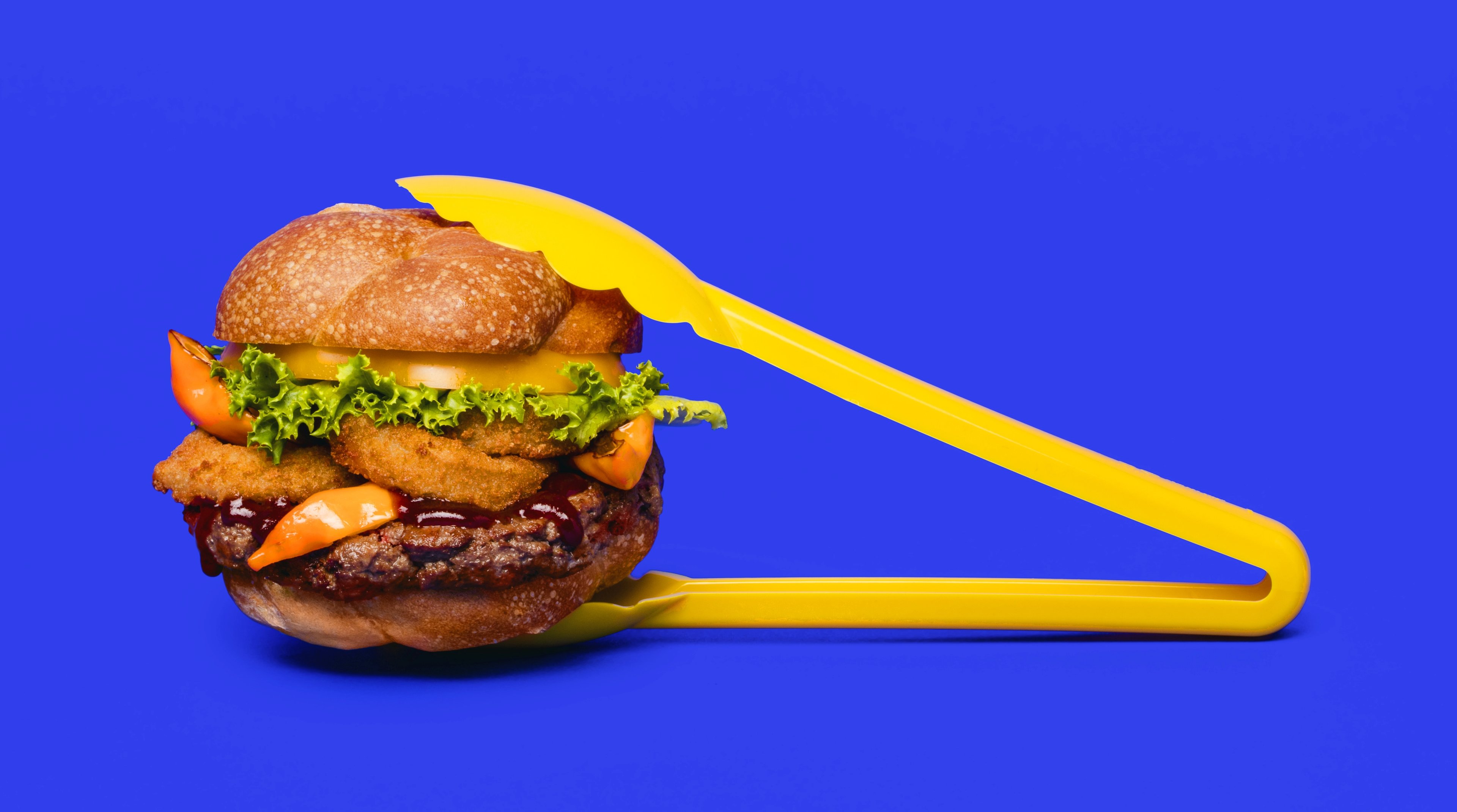 An Impossible Burger held in yellow plastic tongs, with a blue background.