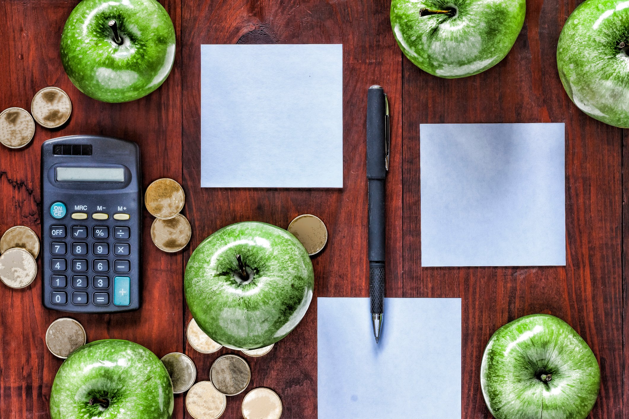 A desk with neatly arranged calculator, pen, papers, gold coins, and green apples.