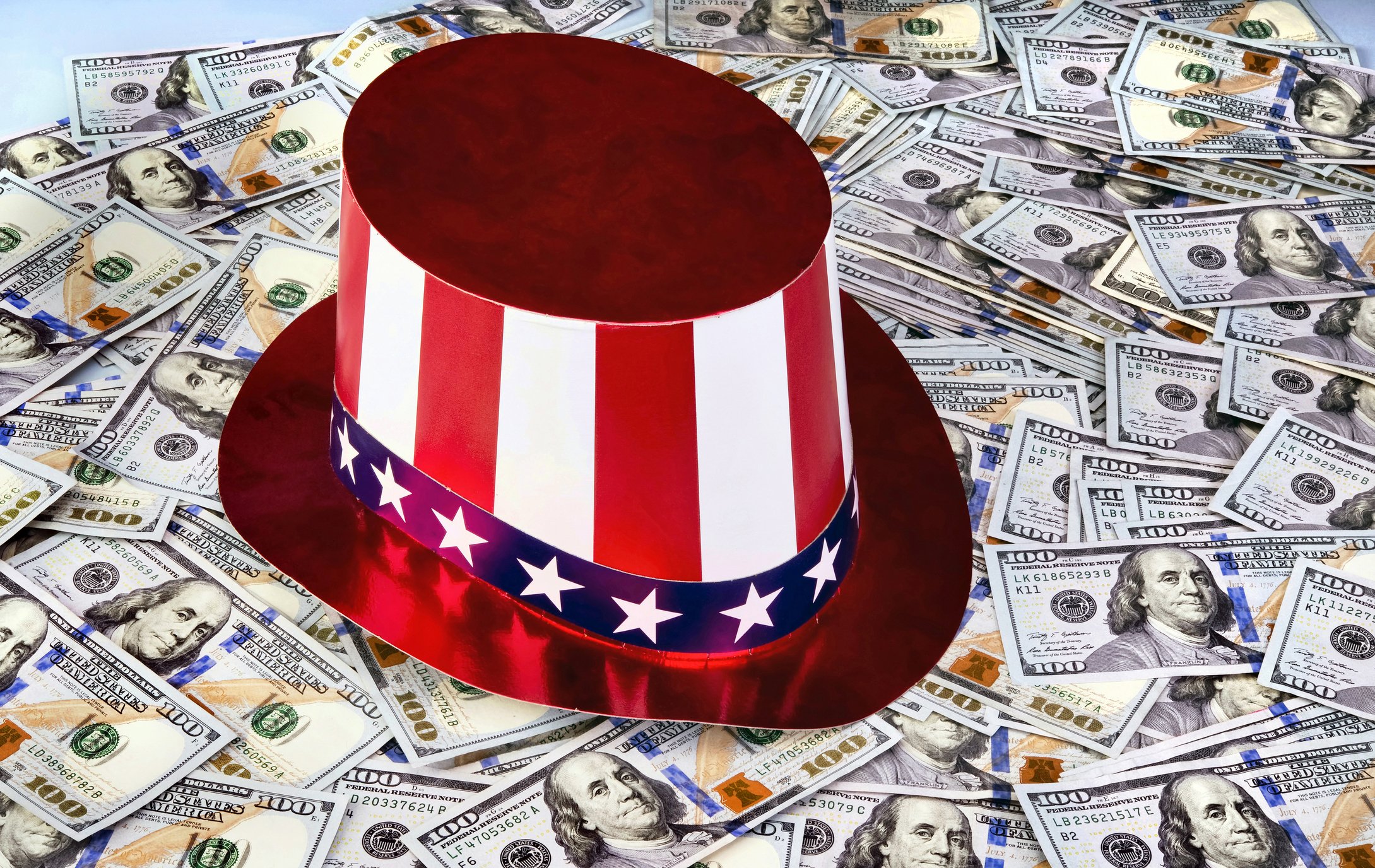 Hat with red, white, and blue stripes and stars sitting on many hundred dollar bills.
