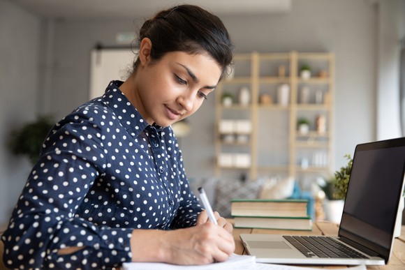 A young woman sitting at her desk, with a laptop in front of her, taking notes on a notebook. 