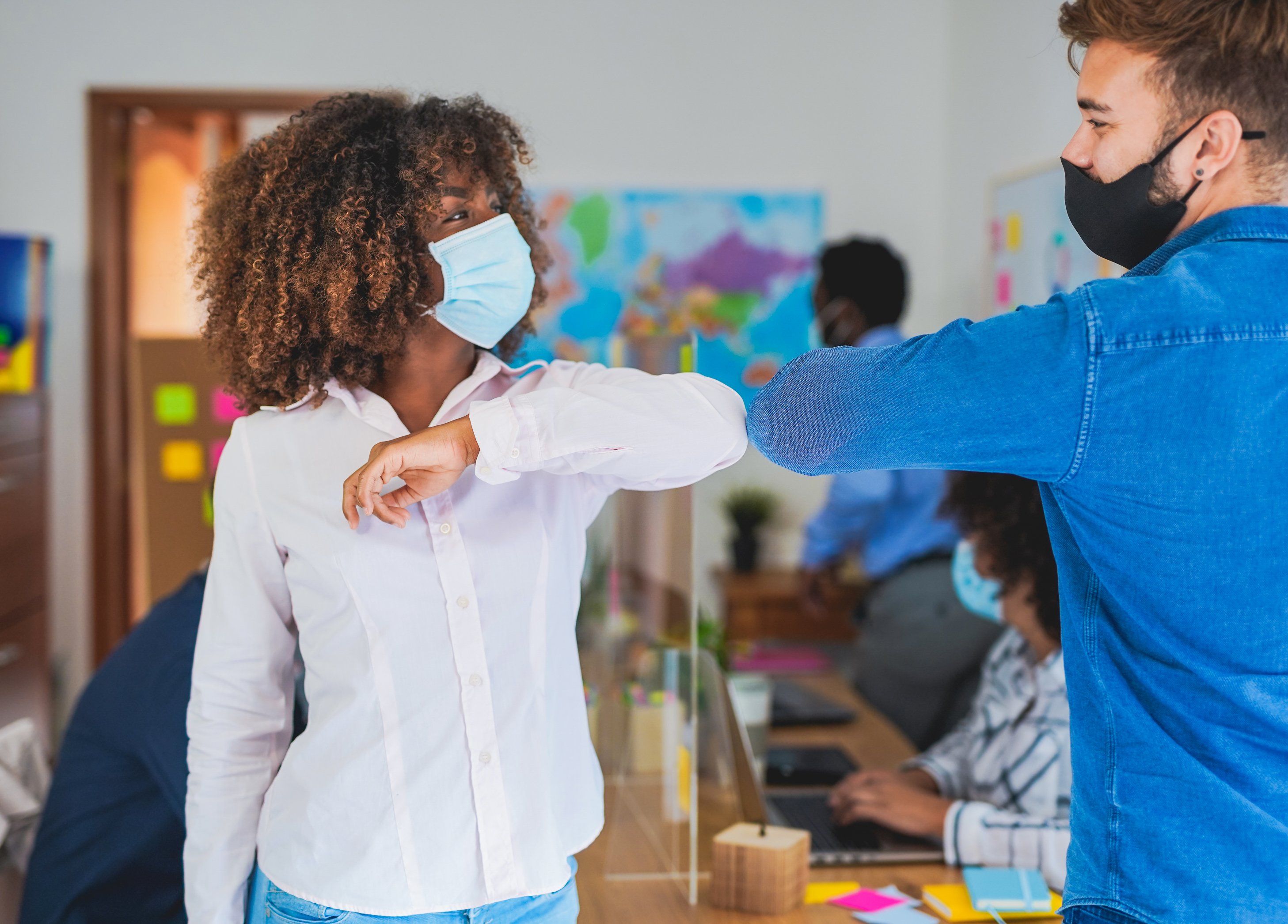 Two young people, both wearing masks, bump elbows in a busy office.
