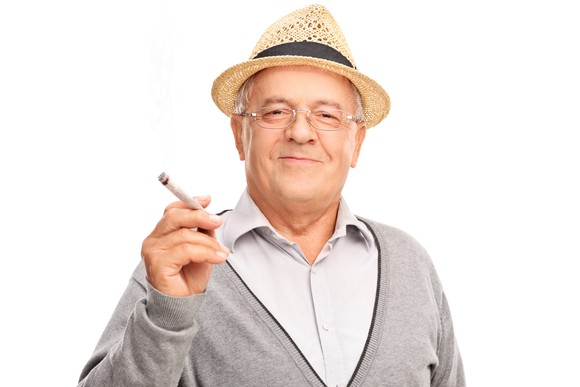 An older man in a hat holds a joint and smiles into camera. 