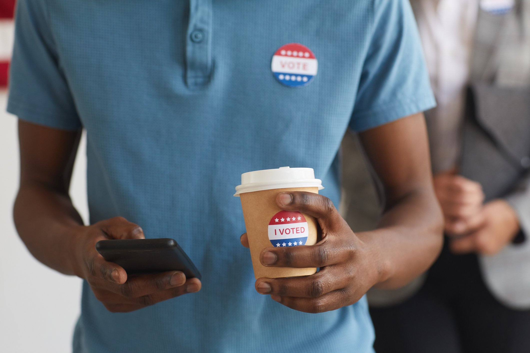 Torso of a man using a smartphone, with election-related stickers on his shirt and on a takeout coffee he's holding.
