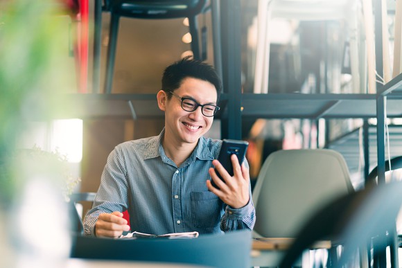A young man in glasses smiles at  his smartphone sitting  at a table. 