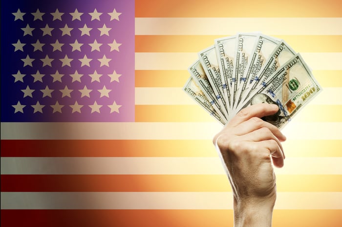 Hand holding $100 bills in front of a U.S. flag