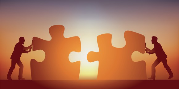 Silhouettes of people pushing two giant jigsaw puzzle pieces together with the sun shining in the background
