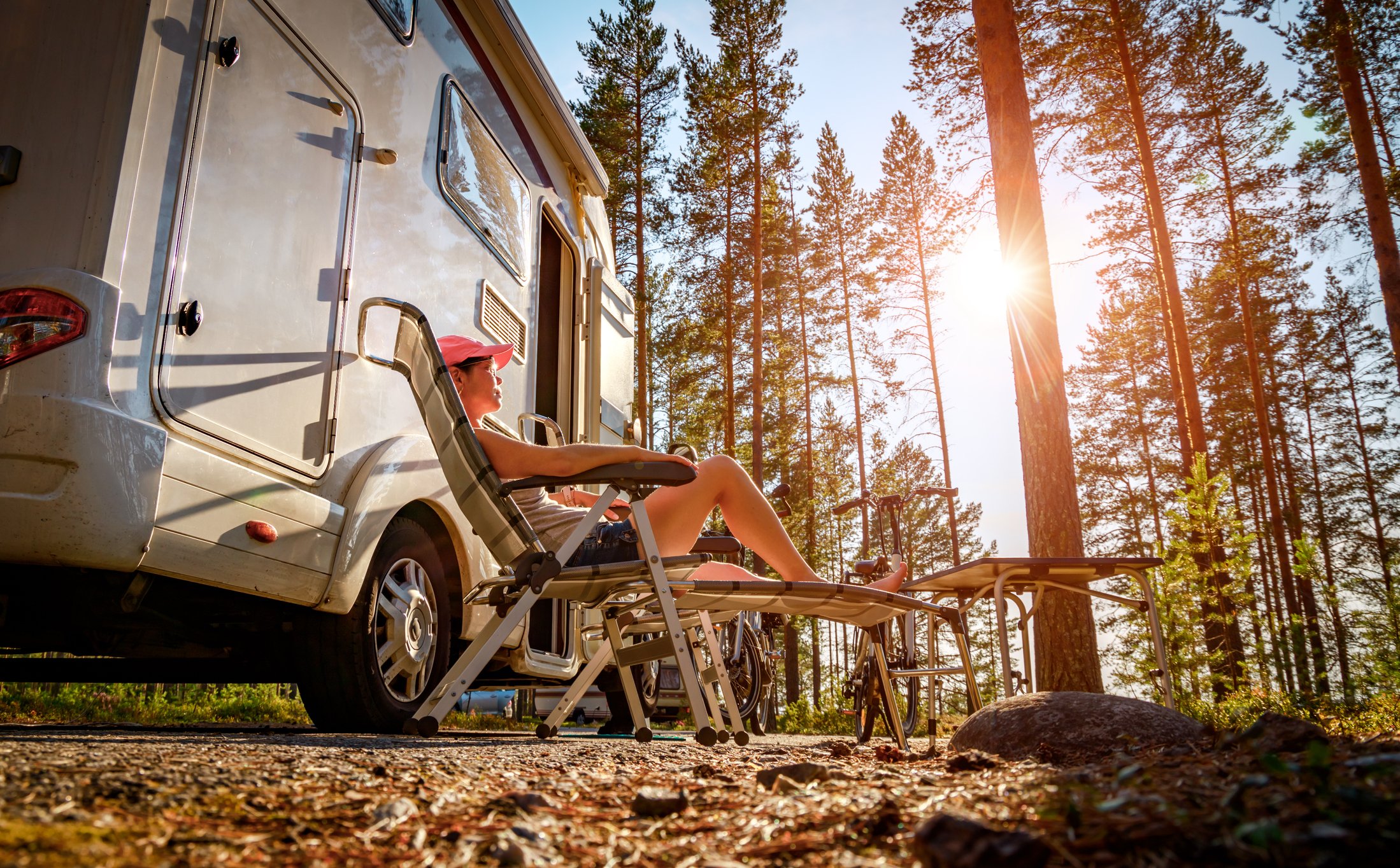 People sitting in lawn chairs outside a motor home in a sunlit pine forest. 