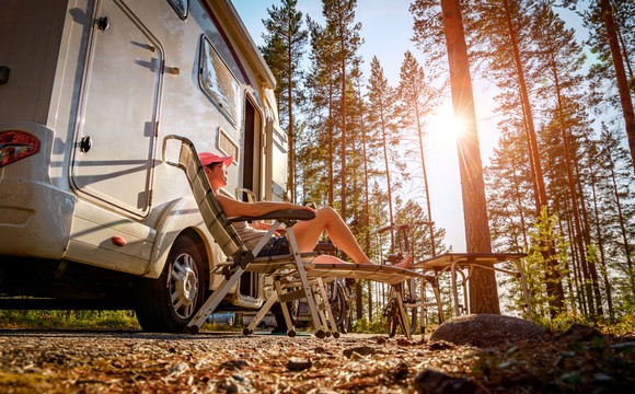 People sitting in lawn chairs outside a motor home in a sunlit pine forest. 