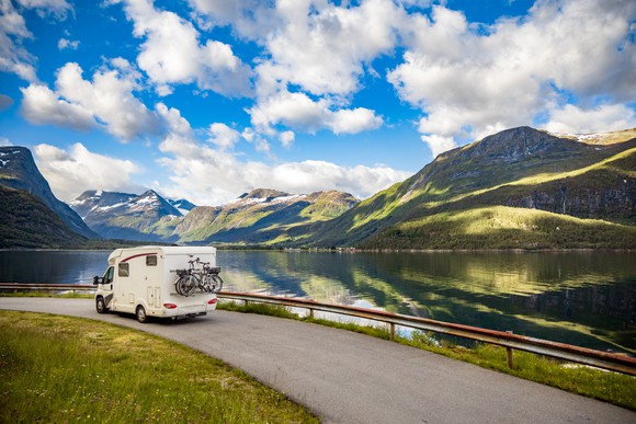 An RV driving along a road next to a lake, with hills and a partly cloudy sky in the background.