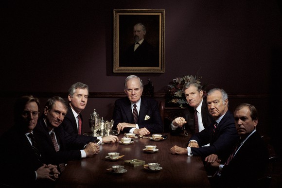 Men in suits sit around a wooden conference table in a dark room. 