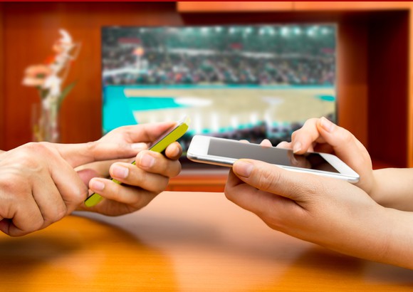 Two people holding phones and a basketball court on a TV in the background.