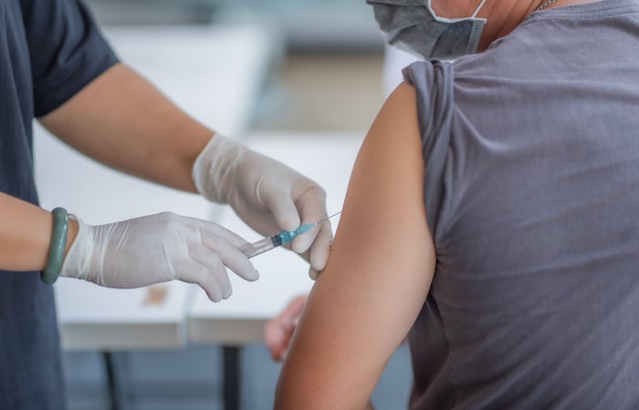 Gloved clinician administers a vaccine to a patient, who wears a mask and a grey t-shirt with sleeve rolled up.