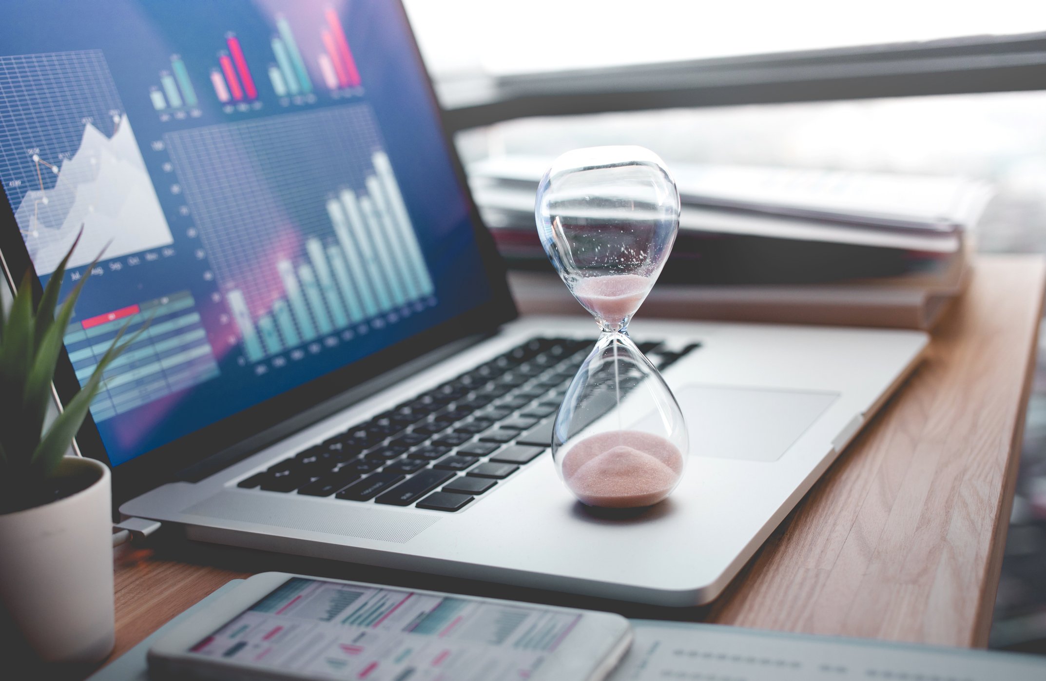 A hourglass rests the palmrest of a laptop displaying charts. 